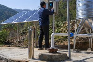 A realistic outdoor installation scene showing a HYBSUN solar powered borehole water pump system working in a remote rural area. A stainless steel HYBSUN submersible pump is displayed beside the wellhead for product reference, while the real pumping setup shows clean white outlet pipes coming from the borehole and connecting to a raised water storage tank and irrigation pipeline. Solar panels are installed nearby to power the system, and a Chinese technician is checking the solar pump controller without showing his face. The scene demonstrates how HYBSUN solar powered water pumps work effectively for off grid water supply, agriculture, livestock, and remote home water pumping applications.