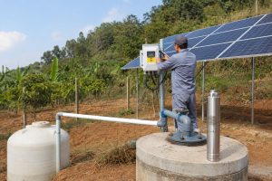 A realistic outdoor installation scene showing a HYBSUN solar pump system set up beside a rural borehole well. A stainless steel deep well solar water pump is displayed upright near the concrete wellhead, while clean new PVC piping runs from the well outlet toward a white water storage tank for irrigation or pond pump system use. A Chinese technician is checking the HYBSUN solar pump controller mounted near the solar panel array, with cables and pipe connections arranged professionally for a working solar water pump installation. The scene includes farmland, green trees, sunlight, solar panels, a water tank, and clean pipework, showing how a HYBSUN solar pump can support off grid water supply, irrigation, and solar powered pond pump applications.