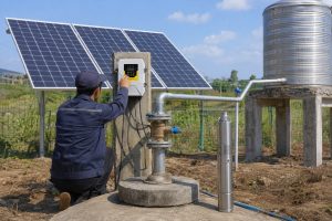 A realistic outdoor HYBSUN solar pump installation scene showing a solar water pump system used for off grid agricultural water supply. A Chinese technician is checking the HYBSUN solar pump controller mounted beside a concrete wellhead, while clean metal pipes connect the borehole outlet to a raised stainless steel water tank. Several solar panels are installed behind the system to power the pump independently from the grid. A stainless steel solar submersible pump is displayed beside the well as a product reference, while the actual working setup shows the water being delivered through a professional pipe network for storage or irrigation. The scene highlights how a HYBSUN solar pump can reduce electricity dependence compared with a normal AC pump, making it suitable for farms, remote water supply projects, pond pump system upgrades, solar powered pond pump applications, and sustainable solar water pump installations.
