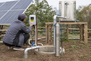 A HYBSUN solar water pump is installed outdoors beside a rural pond in a natural, sunny environment. The scene shows a worker crouched next to the black HYBSUN pond pump, adjusting its components, with clear metallic inlet and outlet pipes directing water from the pond to a connected irrigation system. A solar panel array is positioned nearby, powering the pump, highlighting a sustainable solar powered pond pump setup. Lush greenery and distant hills surround the pump, emphasizing a realistic rural pond pump system installation. The water flows visibly from the pipe, illustrating the pump in active operation, capturing a professional maintenance and repair scenario for a solar water pump.