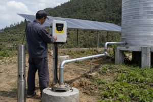 A realistic outdoor installation scene showing a HYBSUN solar water pump system at a remote rural site, with a stainless steel deep well pump displayed beside the borehole while clean metal discharge piping rises from the wellhead and runs toward a large water storage tank and irrigation area. A Chinese technician is checking the HYBSUN solar pump controller mounted near the well, with solar panels installed behind the system to power the pump. The scene shows a professional solar water pump setup for off grid water supply, agricultural irrigation, and remote locations where reliable water access and lower energy costs are important. The clean pipework, practical controller wiring, water tank connection, and cultivated field detail help illustrate how a solar powered pond pump, pond pump system, or deep well HYBSUN solar pump can support daily water delivery in real working conditions.