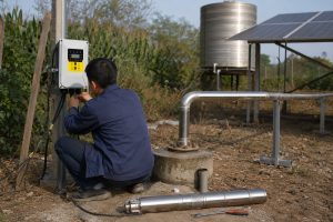 Outdoor rural installation scene showing a HYBSUN solar water pump setup beside a small wellhead. A stainless steel submersible pump with a slim cylindrical body lies on the ground in front of a concrete well base, featuring a perforated lower section and metallic finish. A curved stainless steel pipe with flanged connections emerges from the well, connecting to a horizontal water discharge line leading toward a nearby irrigation system. An AC/DC HYBSUN solar pump controller is mounted on a metal post nearby, displaying a bright yellow panel with buttons and a screen. A Chinese technician wearing a dark blue work jacket and pants is crouched at the controller, adjusting wiring, with only his back and hands visible. The scene includes a metal-frame solar panel array to the right and an elevated cylindrical water tank in the background. Ground is dry with scattered weeds, surrounded by tall crops and greenery, emphasizing a real pond pump system and solar powered pond pump environment. The image realistically depicts a functioning HYBSUN solar pump setup for water supply or irrigation.