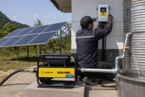 A realistic outdoor installation scene shows a HYBSUN solar pump system set up beside a large stainless steel water tank in a rural area. One Chinese technician, seen from the side and back, is carefully adjusting the wall mounted controller while the single HYBSUN solar water pump sits on a clean concrete base in the foreground. The pump features a sturdy black frame, compact motor body, and visible branded side panel, with new metal inlet and outlet pipes professionally connected for real working use. Several solar panels are installed nearby on a metal support structure, supplying power to the system, while clear water is flowing from the pipe into the tank, showing active operation. Surrounded by open sky, green trees, and a natural hillside setting, the scene presents a durable and practical HYBSUN solar pump solution for long term water supply, and naturally fits searches related to a solar powered pond pump, solar water pump, and efficient pond pump system.