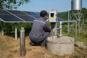 A realistic outdoor HYBSUN solar pump installation scene showing a stainless steel solar well pump displayed upright beside a concrete borehole wellhead, while the actual working pump is positioned underground as part of a professional off grid solar water pump system. A Chinese technician is adjusting the HYBSUN controller mounted near the well, with clean metal outlet pipes, elbows, valves, and fittings connected from the wellhead toward an elevated water tank. Large solar panels behind the installation power the system, creating a practical setup for remote water supply, irrigation, livestock watering, and long term energy savings. The scene highlights how a HYBSUN solar pump can support reliable water independence in rural areas, with visual relevance to solar powered pond pump applications, pond pump system design, and complete solar water pump installations.