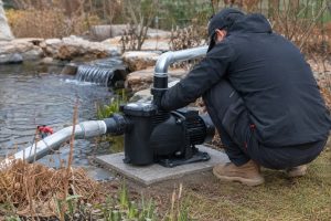 A realistic winter pond maintenance scene showing a black HYBSUN pond pump installed on a concrete equipment pad beside a garden pond. A Chinese technician is crouching with his face hidden while checking the pump and clean metal pipe connections. The pond pump system uses a horizontal intake line from the pond side and a vertical outlet pipe returning water toward a small waterfall, where the water is still circulating in cool weather. The outdoor setting includes natural stones, brown winter plants, leafless shrubs, and a calm backyard pond environment, making the image suitable for explaining whether a solar powered pond pump or solar water pump should stay running through winter. This professional installation scene highlights a practical HYBSUN solar pump solution for pond circulation, winter pond care, and reliable water movement in mild cold climates.