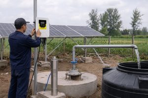 A realistic outdoor HYBSUN solar water pump installation in a rural farmland setting, showing a Chinese technician checking a wall mounted solar pump controller beside a borehole wellhead. A stainless steel deep well pump is displayed neatly beside the concrete well platform, while the working pump remains underground as it would in a real solar water pump system. Clean metal pipes rise from the wellhead through professional fittings and a blue valve, carrying water into a black storage tank with a steady visible flow. Solar panels behind the installation supply power to the controller, creating a practical off grid water solution for irrigation, livestock watering, and remote water supply. The scene naturally illustrates how a HYBSUN solar pump can replace unreliable grid power or fuel driven systems, similar to a solar powered pond pump or pond pump system used for efficient and cost effective water movement.