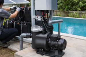 A realistic outdoor installation scene showing a HYBSUN solar pump used as part of a pool pump system beside a residential swimming pool. The black pool pump has a clear strainer lid, compact motor body, and secure base, with clean pipe connections arranged for practical water circulation. A technician is checking the nearby battery storage and controller setup, while solar panels in the background support the solar water pump system for reliable pool operation. This image demonstrates how a battery powered solar water pump can help run a pool pump system with stable energy, similar to solar powered pond pump applications for efficient outdoor water circulation.