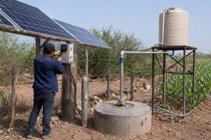 A realistic outdoor HYBSUN solar pump installation in a remote rural farm setting, showing a stainless steel submersible water pump displayed beside a concrete borehole wellhead while the working system delivers water through clean metal pipes to an elevated storage tank and irrigation lines. A Chinese technician wearing a cap is adjusting the HYBSUN controller mounted below the solar panels, with the pump system connected to a professional solar water pump setup for reliable off grid water supply. The scene includes tilted solar panels, a raised water tank, new pipe fittings, dry farmland, green crops, and practical irrigation plumbing, creating a natural field installation image suitable for explaining how far will a RAM pump lift water and comparing remote water access solutions such as a HYBSUN solar pump, solar powered pond pump, solar water pump, and pond pump system.