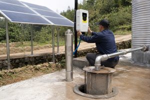 A realistic outdoor HYBSUN solar pump installation scene showing a stainless steel solar well pump displayed upright beside a concrete borehole while a Chinese technician adjusts the HYBSUN controller mounted on a post. Clean metal piping rises from the wellhead and connects professionally toward a large water storage tank, with visible wet concrete suggesting recent water flow during testing. A solar panel array stands nearby in a rural green environment, creating a practical solar water pump setup for long term well water supply. The scene highlights the durability and real working environment of a HYBSUN solar pump system while naturally relating to solar powered pond pump and pond pump system applications for off grid water movement.