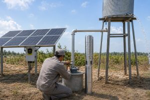 A realistic off grid HYBSUN solar pump installation in a rural outdoor field, showing a stainless steel deep well submersible pump displayed upright beside a concrete borehole wellhead. A Chinese technician is checking the clean metal pipe connections while solar panels power the system through a HYBSUN controller mounted nearby. The discharge pipe runs professionally from the wellhead to an elevated metal water tank, with clear water flowing from the outlet. This practical solar water pump setup demonstrates how a 1 HP pump can support irrigation or water storage in remote areas, offering an efficient alternative to grid powered pumping. The scene also relates to solar powered pond pump applications and pond pump system planning where reliable solar water delivery is needed.