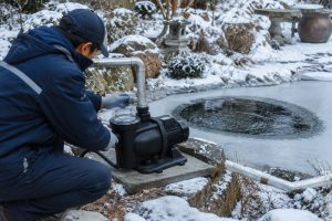 A HYBSUN solar pump is installed beside a partially frozen outdoor pond in a snowy winter garden, showing a realistic cold weather pond pump system for continuous water circulation. The black pond pump sits securely on a stone platform with a transparent strainer lid, horizontal motor housing, and clean metal inlet and outlet pipes connected with professional fittings. A Chinese technician in winter workwear checks the pipe connection while the pond surface remains partly open with visible moving water, helping oxygen flow and reducing the risk of full ice coverage. This real installation scene shows how a solar powered pond pump and solar water pump system can support fish safety and stable pond circulation during winter conditions.