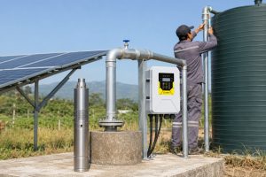 A realistic outdoor HYBSUN solar pump installation on an off grid rural property, showing a stainless steel solar submersible pump displayed beside a concrete borehole wellhead while the actual water line rises from the well through clean galvanized metal pipes. The system includes a HYBSUN solar pump controller mounted on a support frame, solar panels providing power, and a professional pipe connection leading to a large water storage tank for dependable farm and household water supply. A Chinese technician in work clothes is checking the metal pipe near the tank, creating a natural installation scene for a solar water pump system. The image highlights a reliable HYBSUN solar pump solution for deep well water supply, irrigation, and remote properties, while also supporting related solar powered pond pump and pond pump system applications.