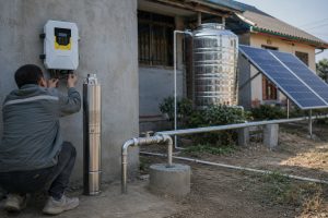A realistic outdoor home water supply installation showing a HYBSUN solar pump system beside a rural house, with one stainless steel solar deep well pump displayed upright next to the wall while the actual borehole setup is connected through clean metal pipes. A Chinese technician is checking the wall mounted solar controller, with the pump linked to a professional wellhead piping system that runs toward a stainless household water tank and nearby solar panels. The scene shows a practical solar water pump installation for improving weak household water pressure and unreliable water supply, with no visible open water flow because the system is connected through sealed pipes. This setup is suitable for home water supply, irrigation support, or similar solar powered pond pump and pond pump system applications where efficient off grid pumping is needed.