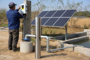 A realistic outdoor HYBSUN solar pump installation scene showing a stainless steel solar water pump displayed beside a concrete borehole wellhead, with clean galvanized metal pipes connected through professional elbows and fittings toward a water storage basin and irrigation line. A Chinese technician is adjusting the HYBSUN solar pump controller mounted on a concrete post, while a solar panel array powers the system in a rural farm setting. The scene highlights a cost effective solar water pump solution for long term water supply, pond pump system applications, and solar powered pond pump installations where reliable performance and lower operating costs matter.