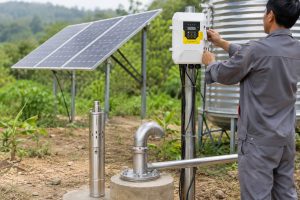 A realistic outdoor HYBSUN solar pump installation scene showing a stainless steel solar water pump displayed beside a borehole wellhead, with clean metal piping connected from the well to a nearby water storage tank and irrigation line. A Chinese technician is adjusting the white HYBSUN solar pump controller mounted on a metal post, while solar panels provide power in the background. The setup highlights an off grid water supply system where voltage choice affects cable size, energy efficiency, and long distance power delivery. The professional installation includes a polished metal discharge pipe, bolted wellhead fittings, organized wiring, and a rural field environment. This image is suitable for explaining the difference between 12V and 24V water pump systems, while also relating to solar water pump applications, HYBSUN solar pump solutions, solar powered pond pump selection, and pond pump system planning for off grid water use.