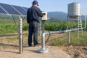 A realistic outdoor HYBSUN solar pump installation shows a stainless steel deep well submersible pump displayed beside a working borehole system, with the actual pump installed underground and a clean metal outlet pipe rising from the wellhead. A Chinese technician is checking the solar pump controller near a ground mounted solar panel array, while galvanized metal pipes, elbows, valves, pipe supports, and a blue connector guide water toward an elevated storage tank for off grid irrigation and rural water supply. The scene presents a powerful solar water pump solution for areas without grid access, offering a stronger and more independent alternative to a small solar powered pond pump or basic pond pump system.