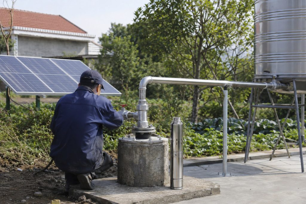 A realistic outdoor HYBSUN solar pump installation scene showing a stainless steel solar water pump displayed beside a concrete borehole wellhead while the working pump system is connected underground. A Chinese technician is inspecting the clean metal piping, with new stainless steel elbows, valves, unions, clamps, and support brackets guiding water from the wellhead toward a raised metal storage tank. Solar panels are installed beside the system to power the off grid water supply, creating a professional and reliable setup for homes, farms, irrigation, and related applications such as a solar powered pond pump or pond pump system when matched with the proper HYBSUN solar pump model.