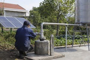 A realistic outdoor HYBSUN solar pump installation scene showing a stainless steel solar water pump displayed beside a concrete borehole wellhead while the working pump system is connected underground. A Chinese technician is inspecting the clean metal piping, with new stainless steel elbows, valves, unions, clamps, and support brackets guiding water from the wellhead toward a raised metal storage tank. Solar panels are installed beside the system to power the off grid water supply, creating a professional and reliable setup for homes, farms, irrigation, and related applications such as a solar powered pond pump or pond pump system when matched with the proper HYBSUN solar pump model.
