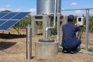 A realistic outdoor HYBSUN solar pump installation scene showing a stainless steel submersible solar water pump displayed beside a concrete borehole wellhead, with clean galvanized metal pipes rising from the well and connecting toward a raised water storage tank. A Chinese technician in blue workwear is adjusting the wall mounted solar pump controller while solar panels provide off grid power in the background. The setup illustrates a reliable HYBSUN solar pump system for farms, irrigation, deep wells, and remote water supply, with professional pipe connections and a practical installation layout. Unlike a small solar powered pond pump or pond pump system, this solar water pump is designed for secure off grid water delivery from a borehole to storage or irrigation lines.