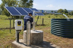 A realistic outdoor HYBSUN solar pump installation scene showing a stainless steel submersible borehole pump displayed beside a concrete wellhead, with clean metal discharge piping rising from the well and directing flowing water into a large storage tank. A HYBSUN solar pump controller is mounted near the well, connected to a solar panel array in the background, while a Chinese technician checks the system from behind. The professional off grid setup highlights reliable solar water pump performance for deep wells, irrigation, water storage, and buyers comparing a solar powered pond pump or pond pump system for cost effective water solutions.