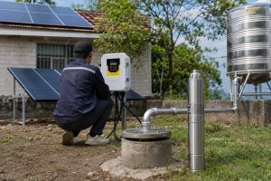A realistic outdoor HYBSUN solar pump installation scene showing a Chinese technician checking a solar water pump controller beside a concrete borehole wellhead in a rural home yard. Clean metal piping comes from the covered well and connects professionally toward a raised stainless steel water tank, while a stainless steel submersible deep well pump is displayed upright next to the installation for product reference. Solar panels are mounted on the house roof and on a ground rack, creating a complete HYBSUN solar pump system for household water supply and irrigation. The scene highlights the practical setup, maintenance access, pipe connections, controller inspection, and real working environment of a solar water pump system, with related applications such as solar powered pond pump and pond pump system installations.