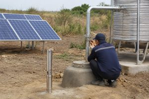 A realistic outdoor HYBSUN solar pump installation scene showing a stainless steel submersible well pump displayed beside a concrete borehole, with clean metal outlet piping rising from the wellhead and connecting to a large stainless steel water tank. A Chinese technician in workwear is checking the pipe fittings near the well, while solar panels power the solar water pump system in the background. The setup illustrates a professional field inspection for water pressure loss and pump reset troubleshooting, with clear details of the borehole, control area, water tank, and irrigation ready plumbing. This HYBSUN solar pump scene is suitable for explaining well pump reset mechanisms and is related to solar powered pond pump and pond pump system applications.