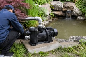 A Chinese technician inspects a single black HYBSUN pond pump installed on a concrete base beside a landscaped garden pond. The pump is connected to clean metal inlet and outlet pipes with professional fittings, while slightly cloudy water and a weak waterfall flow indicate reduced circulation caused by debris buildup. Rocks, aquatic plants, grass, and dense greenery surround the practical outdoor pond pump system. No solar panels are visible, but the installation layout is suitable for a solar powered pond pump or solar water pump configuration using a compatible HYBSUN solar pump power system.