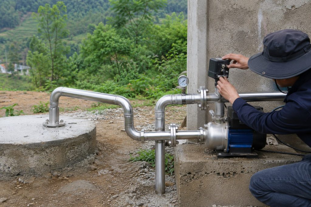 A Chinese technician wearing a dark work hat adjusts the black pressure switch mounted above a blue and stainless steel surface water pump beside a concrete utility building. Clean stainless steel inlet and outlet pipes connect the pump to a covered well opening, while a pressure gauge monitors the system for unstable pressure and frequent pump cycling. No visible water discharge indicates that the equipment is being inspected or tested rather than actively pumping. Green hillside vegetation surrounds the rural installation, creating a realistic outdoor maintenance scene relevant to troubleshooting a HYBSUN solar pump, solar water pump, solar powered pond pump, or similar pond pump system.
