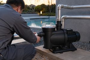 A Chinese technician adjusts a HYBSUN swimming pool pump beside a modern outdoor pool during the early morning off peak electricity period. The black pump features a transparent strainer lid, a compact electric motor and a secure equipment base connected to clean metal inlet and outlet pipes with professional unions and elbow fittings. The calm pool water, landscaped garden and soft dawn lighting create a realistic residential installation scene with no visible leaks or splashing. Although no solar panels are shown, the practical piping layout is also relevant to HYBSUN solar pump applications including a solar water pump solar powered pond pump and professionally installed pond pump system.