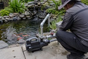 A Chinese technician inspects a compact black pond pump system installed on a concrete platform beside a landscaped 300 gallon koi pond. The surface-mounted pump is connected to clean metal inlet and outlet pipes with professional fittings and a control valve, circulating clear water toward a natural stone waterfall. Green aquatic plants, smooth rocks and colorful koi surround the installation. No solar panels are visible, but the practical setup shows how a HYBSUN solar pump solar water pump or solar powered pond pump can support reliable filtration oxygenation and water circulation in a small garden pond.