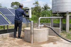 A Chinese technician checks a HYBSUN solar pump controller beside a rural borehole installation powered by several ground mounted solar panels. A slim stainless steel submersible well pump is displayed upright next to the sealed concrete wellhead, while a clean metal discharge pipe rises from the well and connects through professional valves and fittings to an elevated water storage tank. No open water flow is visible, but the connected pipeline shows how the solar water pump transfers groundwater for agricultural storage or irrigation. Surrounded by green crops and trees, the realistic outdoor setup demonstrates the installation layout of a HYBSUN solar pump and helps explain how pumping distance depends on vertical lift, flow rate and pipe resistance. The system differs from a surface solar powered pond pump or pond pump system because the working pump operates below ground inside the well.