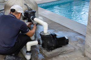 A Chinese pool technician inspects a black HYBSUN swimming pool pump installed on a raised concrete platform beside a residential outdoor pool. The pump is professionally connected to the filtration system with clean intake and return pipes, secure unions, and a multiport valve. Visible ripples across the blue pool water indicate active circulation while the technician adjusts the plumbing controls. No solar panels are present, making this a conventional pool circulation installation rather than a solar powered pond pump, solar water pump, pond pump system, or HYBSUN solar pump setup.