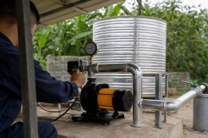 A technician adjusts the automatic pressure controller on a blue HYBSUN surface water pump installed outdoors on a concrete base. The pump is professionally connected to clean stainless steel inlet and outlet pipes, with a visible pressure gauge for monitoring system pressure and water flow. A large metal water storage tank, green vegetation, and a practical utility area appear in the background. No solar panels are visible, but the installation demonstrates a reliable pond pump system that can also support solar water pump applications, including a solar powered pond pump or HYBSUN solar pump setup.