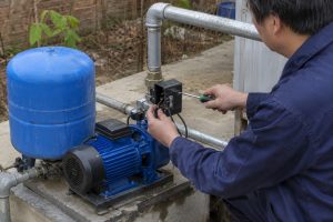 A Chinese technician replaces and adjusts the pressure switch on a blue HYBSUN water pump installed beside a matching pressure tank on a concrete outdoor platform. The pump is connected to clean galvanized metal inlet and outlet pipes with professional threaded fittings and valves, while the open switch housing reveals the electrical terminals being checked with a screwdriver. The dry pipework indicates that the system has been powered off and drained before maintenance. No solar panels are visible, but the practical setup is relevant to pressure control and servicing for a HYBSUN solar pump solar water pump solar powered pond pump or similar pond pump system.
