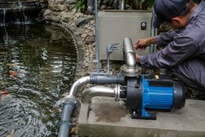 A Chinese technician inspects a blue and black HYBSUN pond pump system installed on a concrete platform beside a landscaped koi pond. Clean stainless steel inlet and outlet pipes connect the pump to the pond circulation system, while a nearby control box supports safe operation. Fish are visible in the water as a small waterfall provides limited aeration. No solar panels are shown, but the installation can represent a solar powered pond pump or solar water pump setup designed to restore circulation and protect fish during a pump failure.