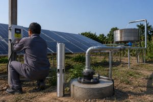 A realistic outdoor HYBSUN solar pump installation scene showing a stainless steel submersible solar water pump displayed beside a borehole wellhead while a technician checks the HYBSUN controller mounted near the solar panels. Clean galvanized metal pipes rise from the well opening and connect professionally to an elevated water tank, with water flowing from the discharge pipe into the tank. The setup includes solar panels, secure pipe fittings, a concrete well base, and a rural irrigation environment, giving a clear view of how a modern solar water pump system can reduce installation complexity compared with traditional pump setups. This scene is also relevant for users comparing solar powered pond pump installation, pond pump system layouts, and HYBSUN solar pump solutions for agricultural water supply.