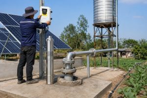 A realistic outdoor installation scene showing a HYBSUN solar pump system for reliable water supply in a rural irrigation area. A Chinese technician is adjusting the HYBSUN controller beside a borehole wellhead, while a stainless steel submersible pump is displayed next to the installation for product reference. Clean metal pipes connect the well outlet to an elevated water tank and continue toward drip irrigation lines in the field. Solar panels are installed nearby to power the system, making it suitable for areas with unstable grid electricity. The scene highlights a professional solar water pump setup that can support irrigation, farm water storage, and applications similar to a solar powered pond pump or pond pump system.
