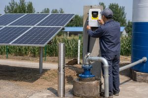 A HYBSUN solar pump installation scene showing a stainless steel submersible well pump displayed beside a concrete borehole, with clean metal outlet piping connected from the wellhead to a large water storage tank. A Chinese technician in work uniform is adjusting the wall mounted HYBSUN solar pump controller while nearby solar panels supply power to the system. The setup shows a realistic solar water pump installation for reliable well water delivery, with professional pipe fittings, electrical conduit, and a rural outdoor environment suitable for irrigation, livestock water supply, or a pond pump system. This image also represents how a solar powered pond pump or HYBSUN solar pump system should be installed with proper plumbing, controller placement, and solar panel connection.