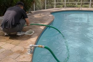 A Chinese pool maintenance technician uses a green garden hose to create a siphon and drain water from a residential swimming pool without relying on the installed HYBSUN pump. The black ground mounted pump remains connected to clean metal inlet and outlet pipes on a concrete equipment platform while water flows steadily through the hose toward a nearby drain. The realistic outdoor setting includes clear pool water, a stone deck, green lawn and landscaped garden. No solar panels are visible, as this manual drainage method is different from a solar powered pond pump solar water pump pond pump system or HYBSUN solar pump installation.