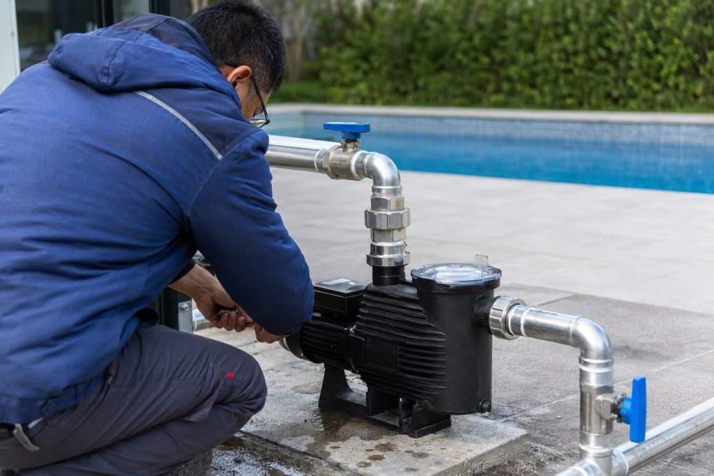 A Chinese pool technician crouches beside a single black HYBSUN variable speed pool pump installed on a concrete equipment pad next to a clean outdoor swimming pool. The pump features a transparent strainer lid and is professionally connected to new stainless steel inlet and outlet pipes with secure unions elbows and blue isolation valves. The technician is adjusting the motor area while the wet surface around the pump suggests recent testing or active water circulation. Clear blue pool water landscaped hedges and a modern residential setting create a realistic maintenance scene. No solar panels are visible because this is a mains powered pool circulation system rather than a solar powered pond pump or solar water pump installation. The image clearly demonstrates how a variable speed pump can support continuous low speed filtration efficient chemical distribution and lower energy use. It is also useful for customers comparing HYBSUN pool equipment with a HYBSUN solar pump or pond pump system.
