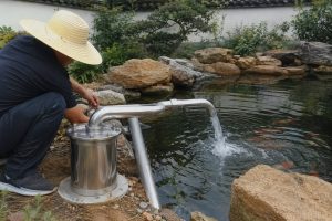 A technician wearing a straw hat checks a cylindrical stainless steel HYBSUN pond pump installed beside a landscaped garden pond. Clean metal inlet and outlet pipes form a professional above ground connection, with a strong stream of clear water flowing from the curved discharge pipe into the pond to improve circulation and oxygen levels. Natural rocks, ornamental plants, rippling water and visible koi create a realistic outdoor installation scene. No solar panels are shown, although this type of pond pump system may be compared with a solar powered pond pump, solar water pump or HYBSUN solar pump for energy efficient pond circulation.