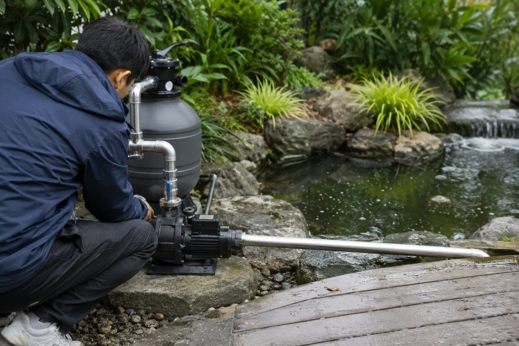 A Chinese technician inspects a compact black HYBSUN garden pond pump installed beside a landscaped backyard pond with rocks, dense green plants, and a flowing waterfall. Clean metal inlet and outlet pipes connect the pump to the pond circulation and filtration system, creating steady water movement that helps prevent stagnant water and supports healthy fish and aquatic plants. Although no solar panels are visible in this installation, the setup demonstrates how a pond pump system, solar powered pond pump, solar water pump, or HYBSUN solar pump can provide reliable circulation for a clear and healthy garden pond.
