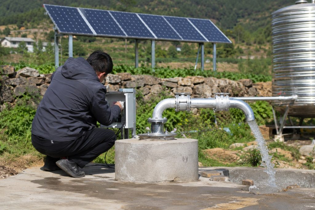 A Chinese technician inspects a HYBSUN solar water pump installation at a rural well site, with a sealed concrete wellhead connected to clean stainless steel outlet pipes leading toward a raised metal water tank. Solar panels provide renewable power for the system while the worker checks the professional pipe fittings and valves. The dry outdoor setting, irrigation land and practical equipment layout show how a HYBSUN solar pump can supply water for farming, storage tanks or a pond pump system, including applications similar to a solar powered pond pump.