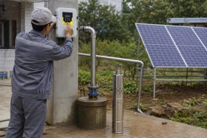 A realistic outdoor home water supply installation scene showing a HYBSUN solar pump system being inspected by a technician beside a residential well. A stainless steel submersible solar water pump is displayed upright next to the wellhead, while the actual working setup uses clean metal piping rising from the borehole and connecting through a professional pipe route for household water delivery. A HYBSUN solar pump controller is mounted on a concrete wall, with a Chinese technician adjusting the control panel during system commissioning. Solar panels stand in the background near a garden area, supplying power to the solar water pump setup. The scene shows a practical alternative to a solar powered pond pump or pond pump system, highlighting reliable off grid water pumping for home use, emergency replacement, irrigation, and daily water supply.