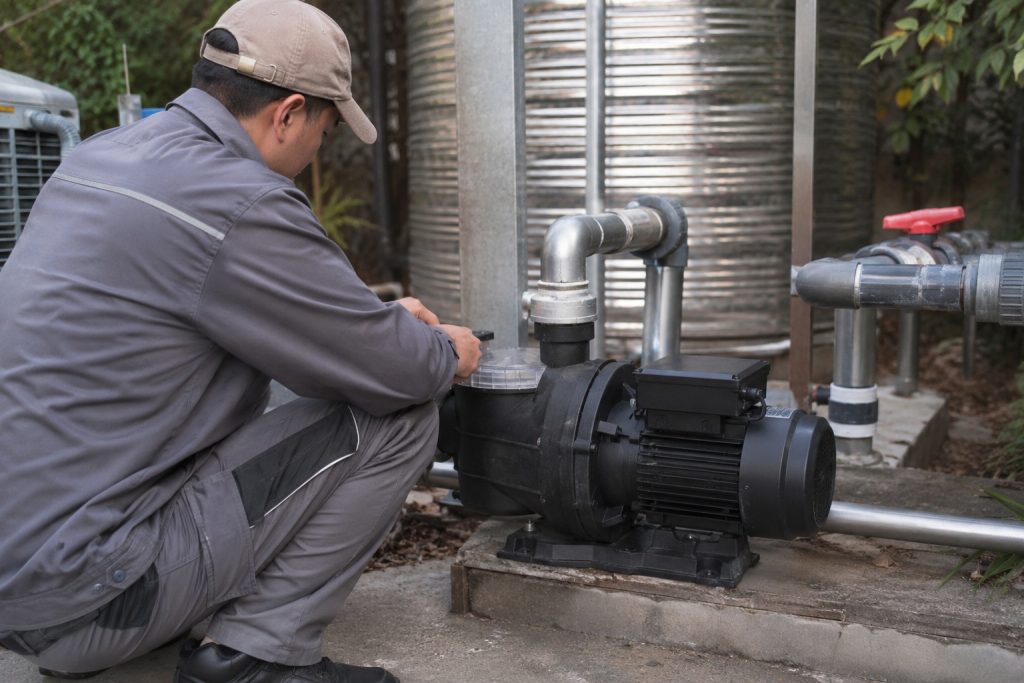A Chinese pool technician inspects a single black variable speed inground pool pump installed on a concrete equipment platform in an outdoor utility area. The pump features a clear strainer lid, a compact motor housing, and professionally connected clean metal inlet and outlet pipes leading toward the pool filtration system and nearby stainless steel water tanks. The secure pipe fittings and valves indicate a functional circulation setup, although no visible water flow is shown. Trees and natural landscaping surround the installation, creating a realistic residential pool equipment scene. No solar panels are present, so this is a conventional pool pump installation rather than a solar powered pond pump or solar water pump system, though HYBSUN solar pump equipment can also support suitable pond pump system applications.