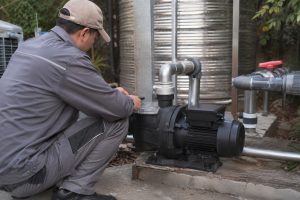 A Chinese pool technician inspects a single black variable speed inground pool pump installed on a concrete equipment platform in an outdoor utility area. The pump features a clear strainer lid, a compact motor housing, and professionally connected clean metal inlet and outlet pipes leading toward the pool filtration system and nearby stainless steel water tanks. The secure pipe fittings and valves indicate a functional circulation setup, although no visible water flow is shown. Trees and natural landscaping surround the installation, creating a realistic residential pool equipment scene. No solar panels are present, so this is a conventional pool pump installation rather than a solar powered pond pump or solar water pump system, though HYBSUN solar pump equipment can also support suitable pond pump system applications.