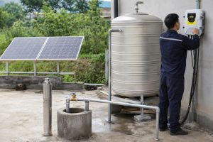 A realistic outdoor home water supply installation featuring a HYBSUN solar pump system with a stainless steel submersible water pump displayed beside a concrete wellhead, clean metal outlet piping connected to a household storage tank, and solar panels powering the system in the background. A Chinese technician is adjusting the wall mounted controller while the new pipework, valve connection, and well outlet show a professional setup designed for stable home water pressure. This scene illustrates how a solar water pump can be used for residential well water supply, garden irrigation, or a pond pump system, offering an efficient alternative to a traditional solar powered pond pump for homes that need reliable off grid water movement.