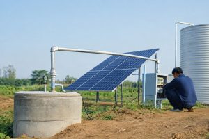 A Chinese technician adjusts the wellhead connections of a HYBSUN solar pump system in a rural field, where a deep well submersible pump operates below ground and lifts water through clean metal piping into a concrete storage tank. A steady stream of water flows from the elevated outlet while large solar panels power the solar water pump through a nearby outdoor controller cabinet. The realistic installation includes secure pipe fittings, electrical cables, dry farmland and distant green hills, showing an off grid water supply arrangement that uses similar solar technology to a solar powered pond pump or pond pump system.