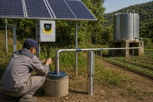 A realistic outdoor installation scene showing a HYBSUN solar pump system on an off grid rural property, with solar panels mounted above a HYBSUN solar pump controller and clean metal piping connected from a borehole wellhead to a raised water tank and irrigation lines. A Chinese technician is checking the pipe connection beside the well, while a stainless steel submersible water pump is displayed upright near the installation area. The setup demonstrates how a solar water pump can provide reliable water for farms, gardens, remote homes, and pond pump system applications without relying on grid power. This professional HYBSUN solar pump scene also reflects the practical use of an aftermarket solar powered pond pump solution for dependable off grid water supply.